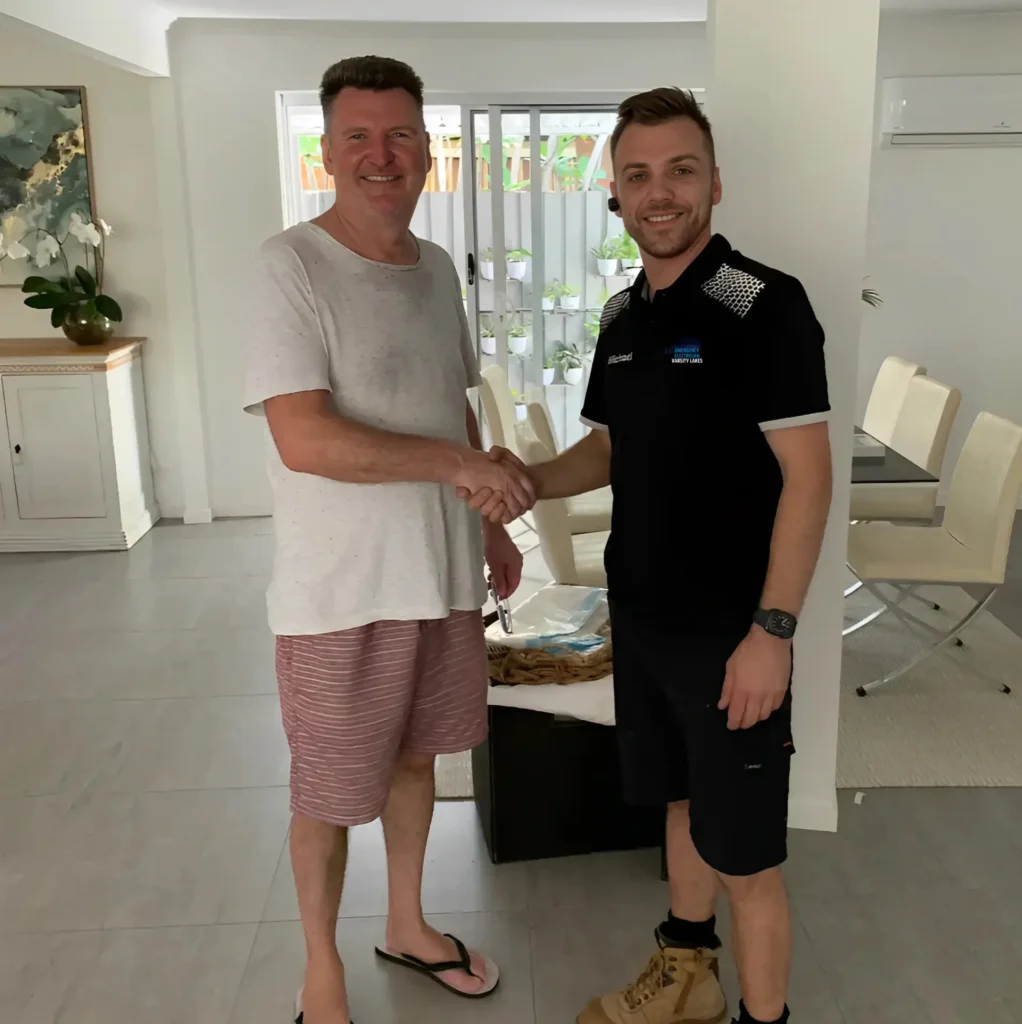 Two men shaking hands in a modern living room, one wearing a casual white shirt and shorts, and the other in a uniform